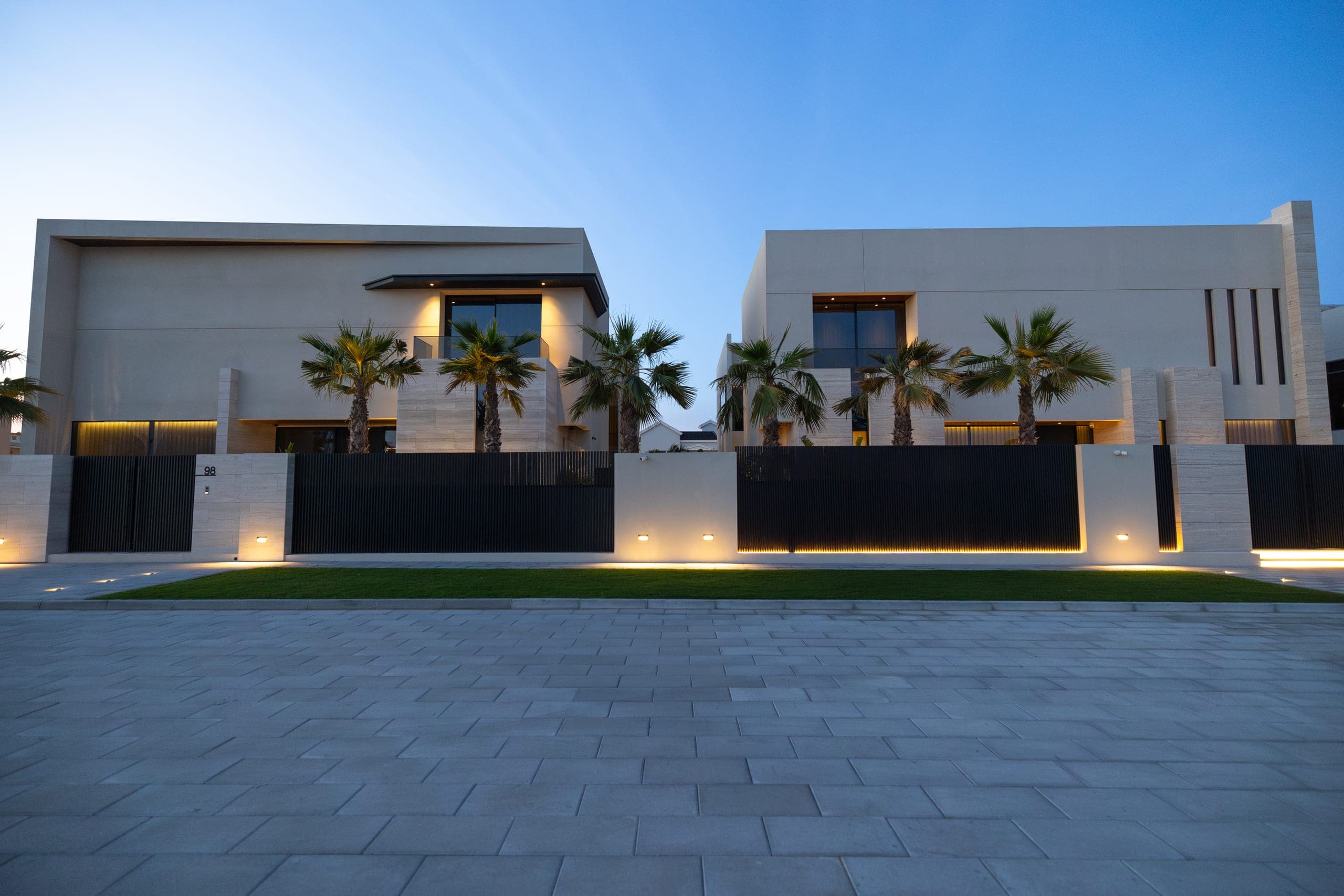 Two adjoining private villas by LKYOOB at dusk — travertine and rendered volumes above a slatted boundary screen, washed palms and a lit forecourt under a deep blue sky